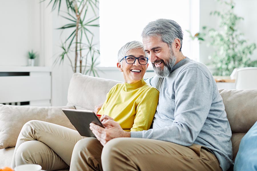 Client Center - Senior Couple Laugh as Their Use a Tablet on Their Couch in Their White Living Room Filled With Plants